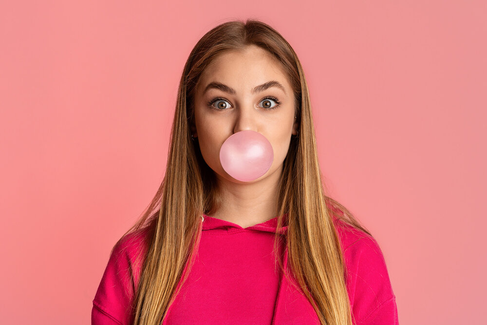 Young woman in a bright pink top blowing a pink bubble gum bubble against a matching pink background.  Image 2 (Right)