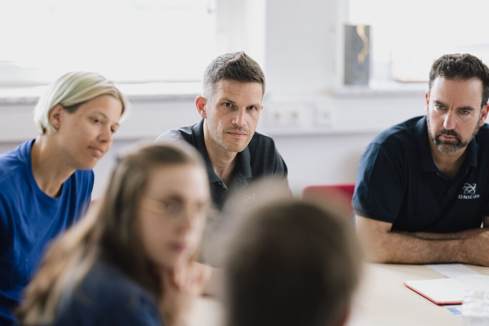 Three adults sit at a table, looking attentively toward someone off-camera. A woman with blonde hair and two men in dark polo shirts are in focus, while two other people in the foreground are blurred.