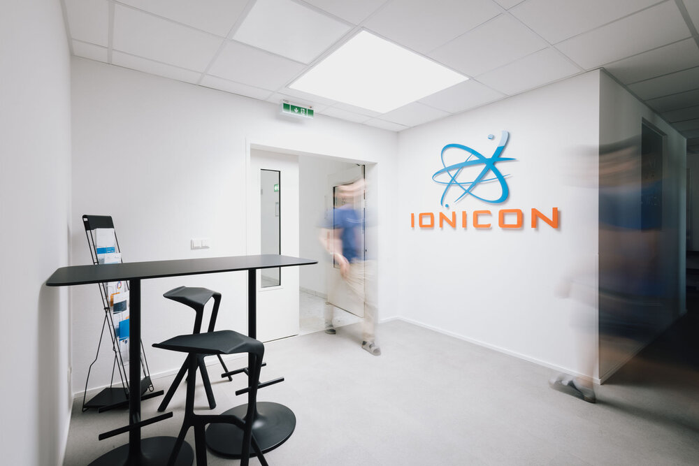 A modern, bright office interior featuring a high black table with two stools. On the white wall, a blue and orange logo reads IONICON with an atomic symbol. A blurred figure of a person is walking through a doorway in the background.
