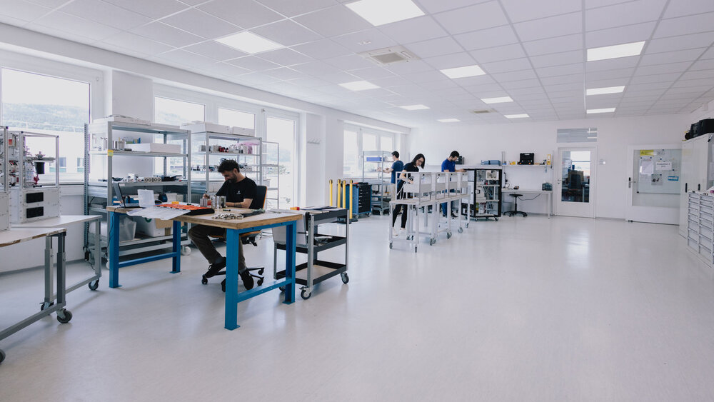 Three technicians work in a bright, spacious, and clean production setting of IONICON. They are positioned at various workstations equipped with tools, shelving, and equipment, focusing on technical tasks in a professional environment.