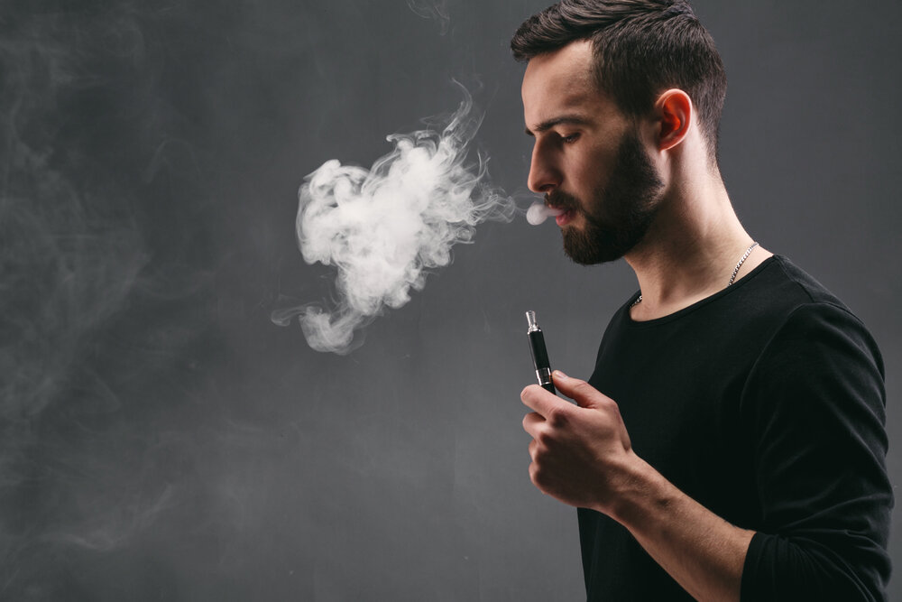 A bearded man in a black shirt exhaling a cloud of vapor from an e-cigarette.